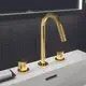 3-Loch Waschtischarmatur in gebürstetem Gold von Ideal Standard mit einem Bogenhahn und zwei Bedienelementen auf einem weißen Waschbecken vor schwarz gefliester Wand.